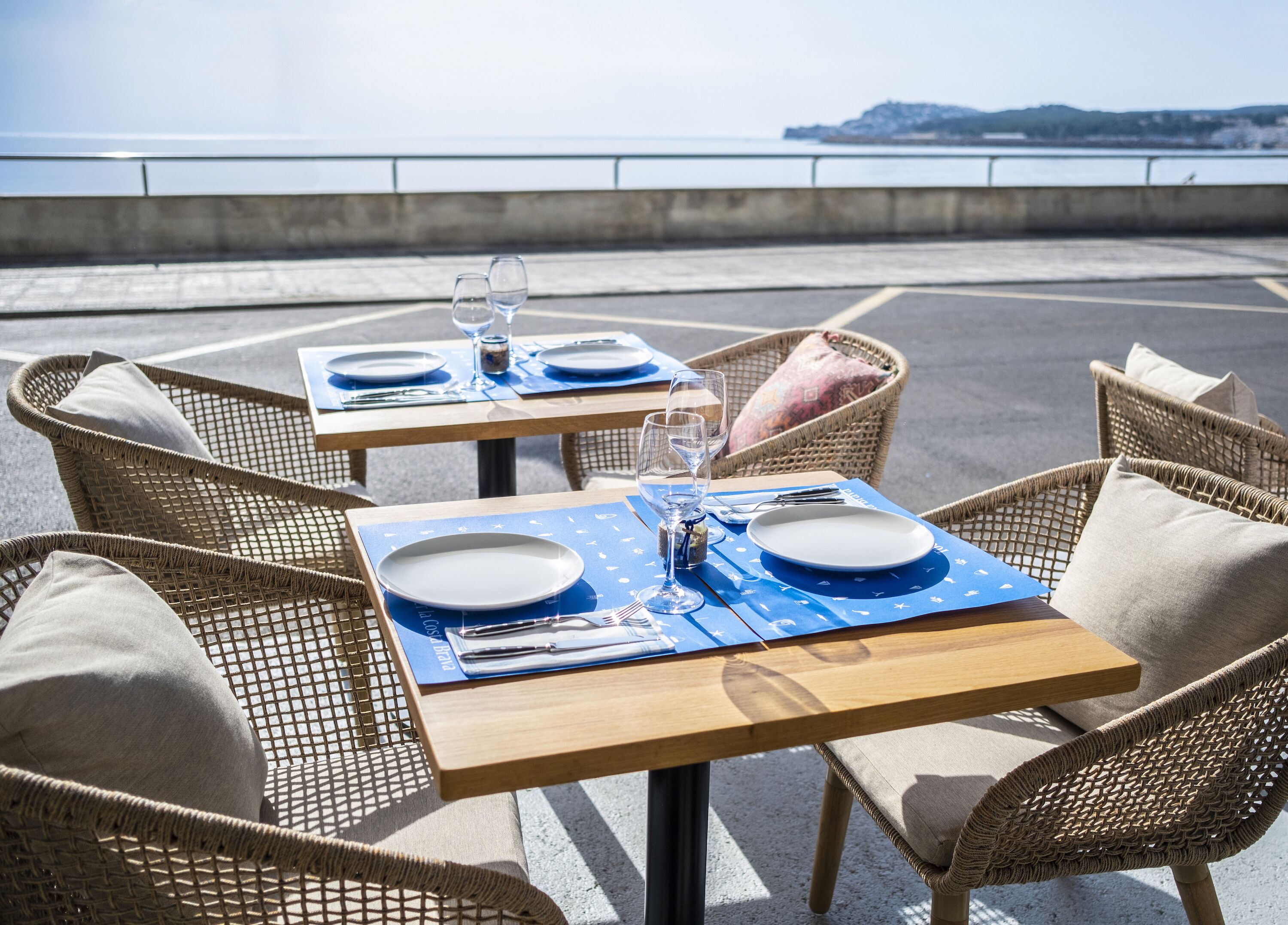 A dining table set with plates, cutlery, and glasses, surrounded by wicker chairs with cushions, overlooking a coastal view.
