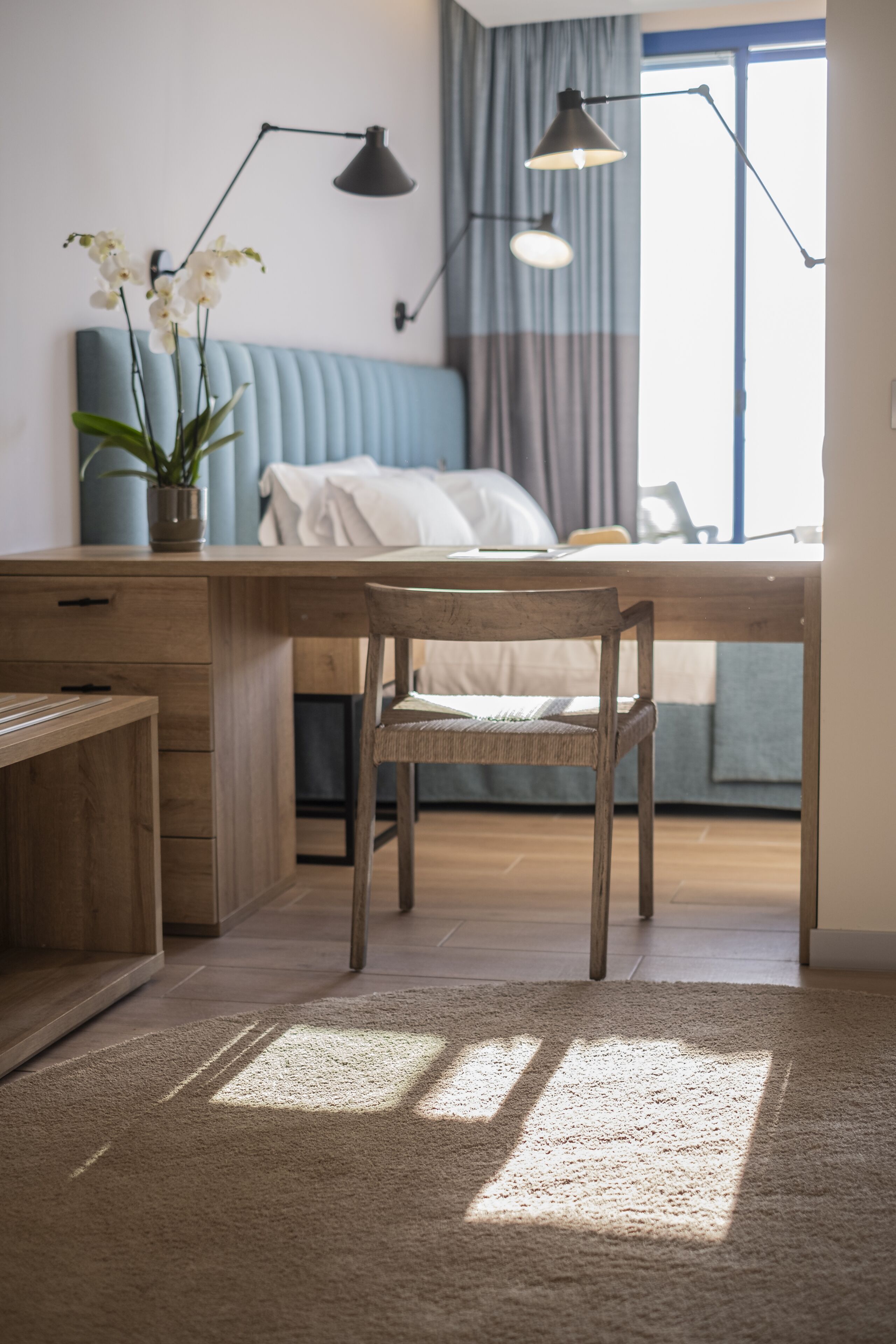 Un dormitorio moderno con un escritorio de madera, una silla, una cama con cabecera azul y una ventana con cortinas.