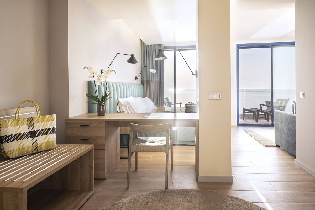 A modern bedroom with a wooden desk, a chair, a bedside table, and a large window offering a view of the outdoors.