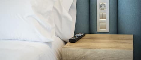 A neatly made bed with white linens, a wooden nightstand with a remote control, and a wall-mounted electrical outlet.