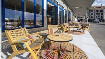 An outdoor seating area with wicker furniture and a patterned rug, in front of a building with large windows and blue trim.