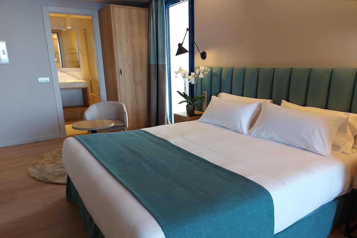 A modern hotel room with a large bed, a small round table, a chair, and a view of the outside.