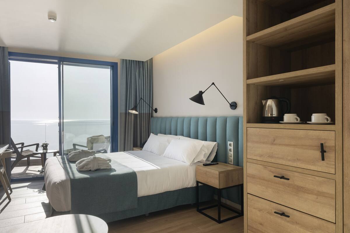 A modern hotel room with a large bed, a wooden wardrobe, a desk, and a view of the sea.