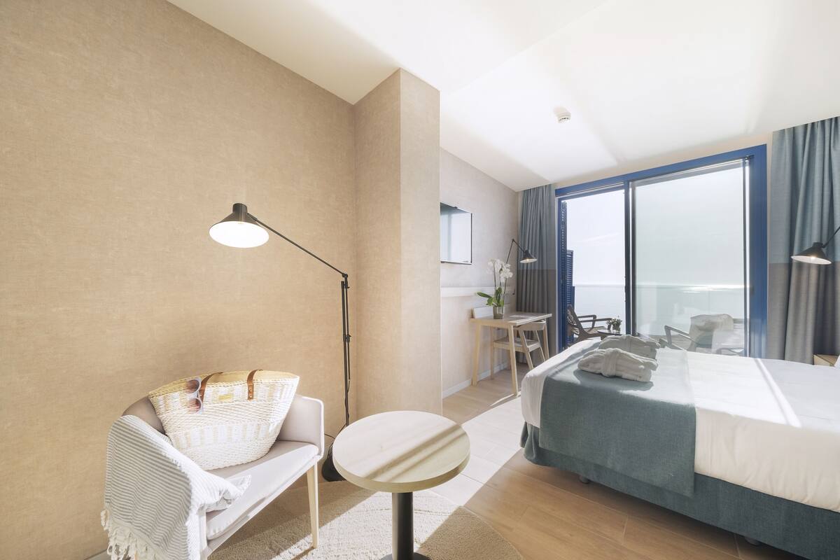 A hotel room with a bed, a round table, a chair, a desk, a lamp, and a view of the city.