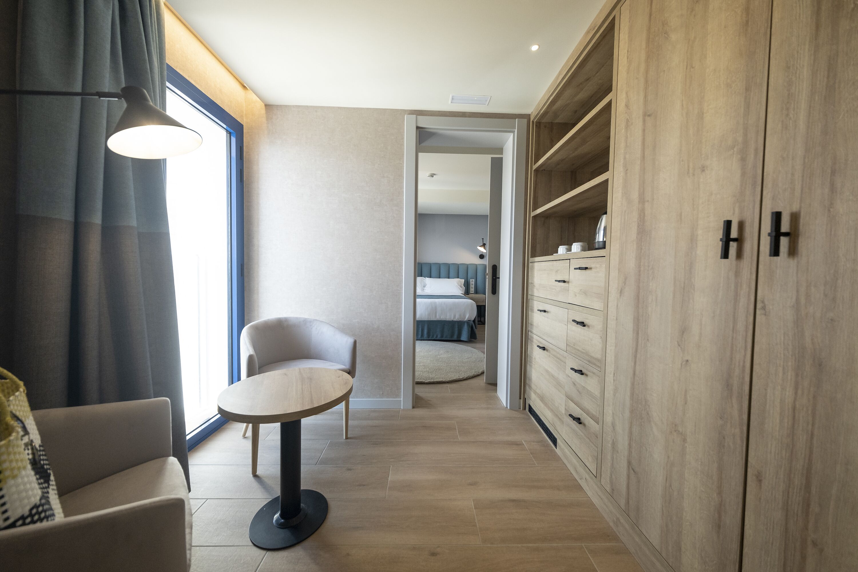 A modern hotel room with a round table, a sofa, a wardrobe, and a bed in the background.