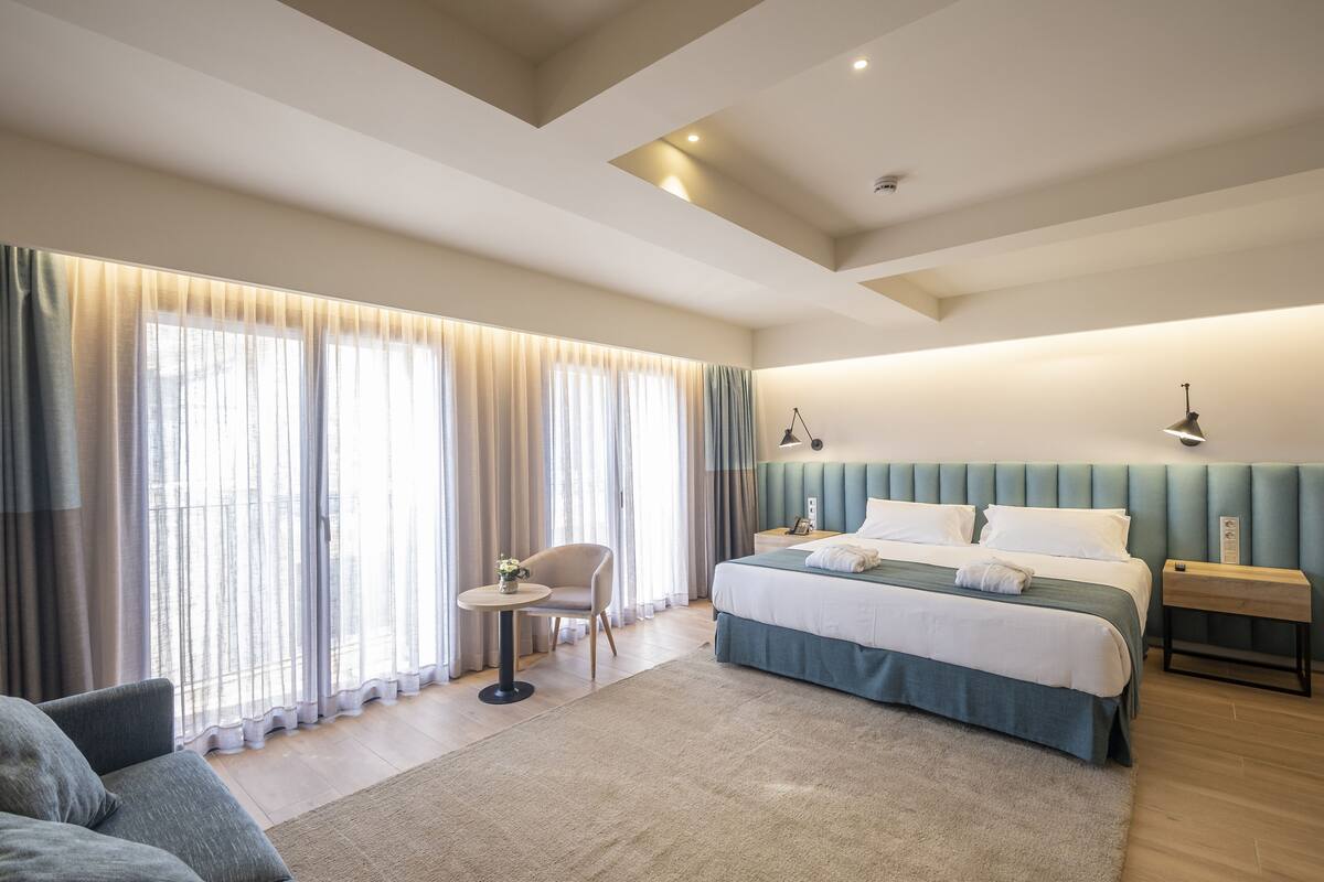 A modern hotel room with a large bed, a seating area with a chair, a small table, and a bedside table with a lamp.
