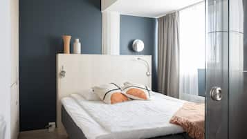 Una cama bien tendida con sábanas blancas y dos almohadones decorativos, una mesita de noche con un florero y un espejo, y una ventana con cortinas.