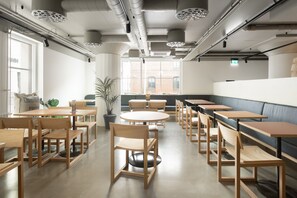 A modern dining area with wooden tables and chairs, a large window with a view of a brick building, and a green plant in the corner. - Noli Katajanokka (Helsinki)