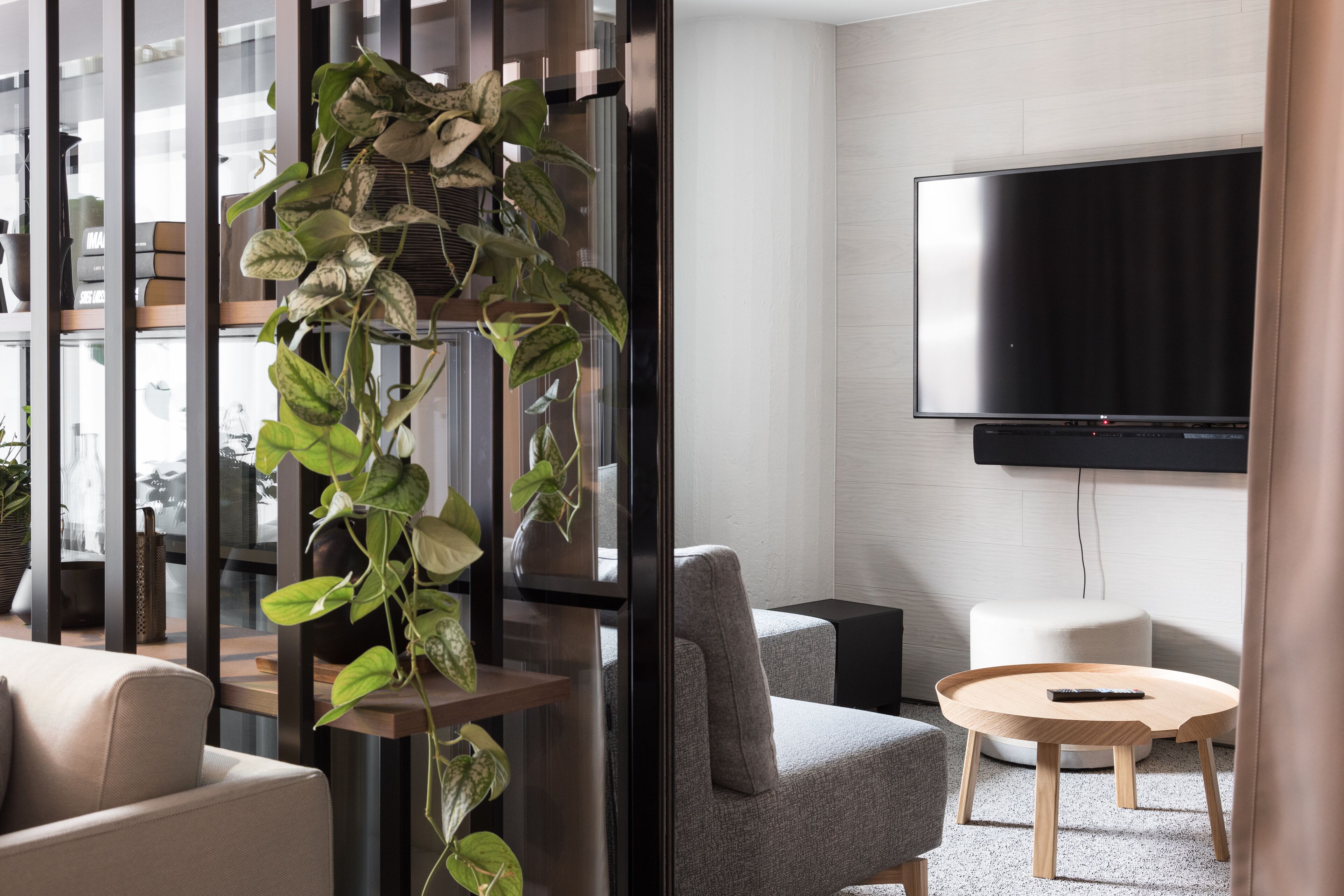 A modern living room with a grey sofa, a round wooden coffee table, a flat-screen TV mounted on the wall, and a large potted plant.