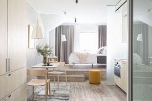 A modern hotel room with a bed, a desk, a chair, a small table, a plant, and a kitchen area. - Noli Katajanokka (Helsinki)