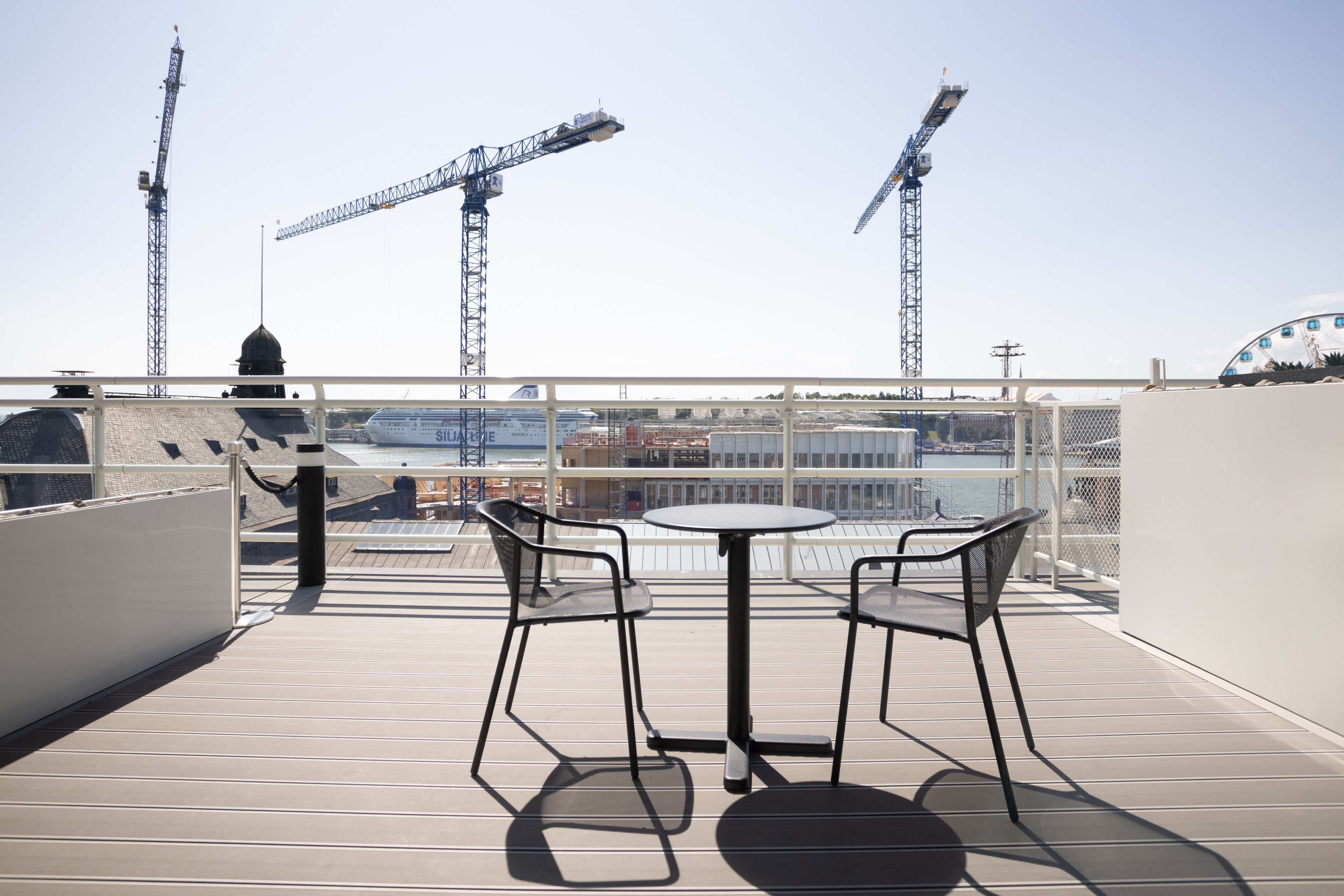 A rooftop terrace with two tables and chairs, overlooking a construction site with cranes.