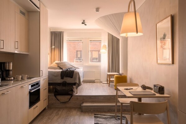 A compact hotel room with a kitchenette, a bed, a small table with a chair, and a window with curtains. - Noli Katajanokka (Helsinki)