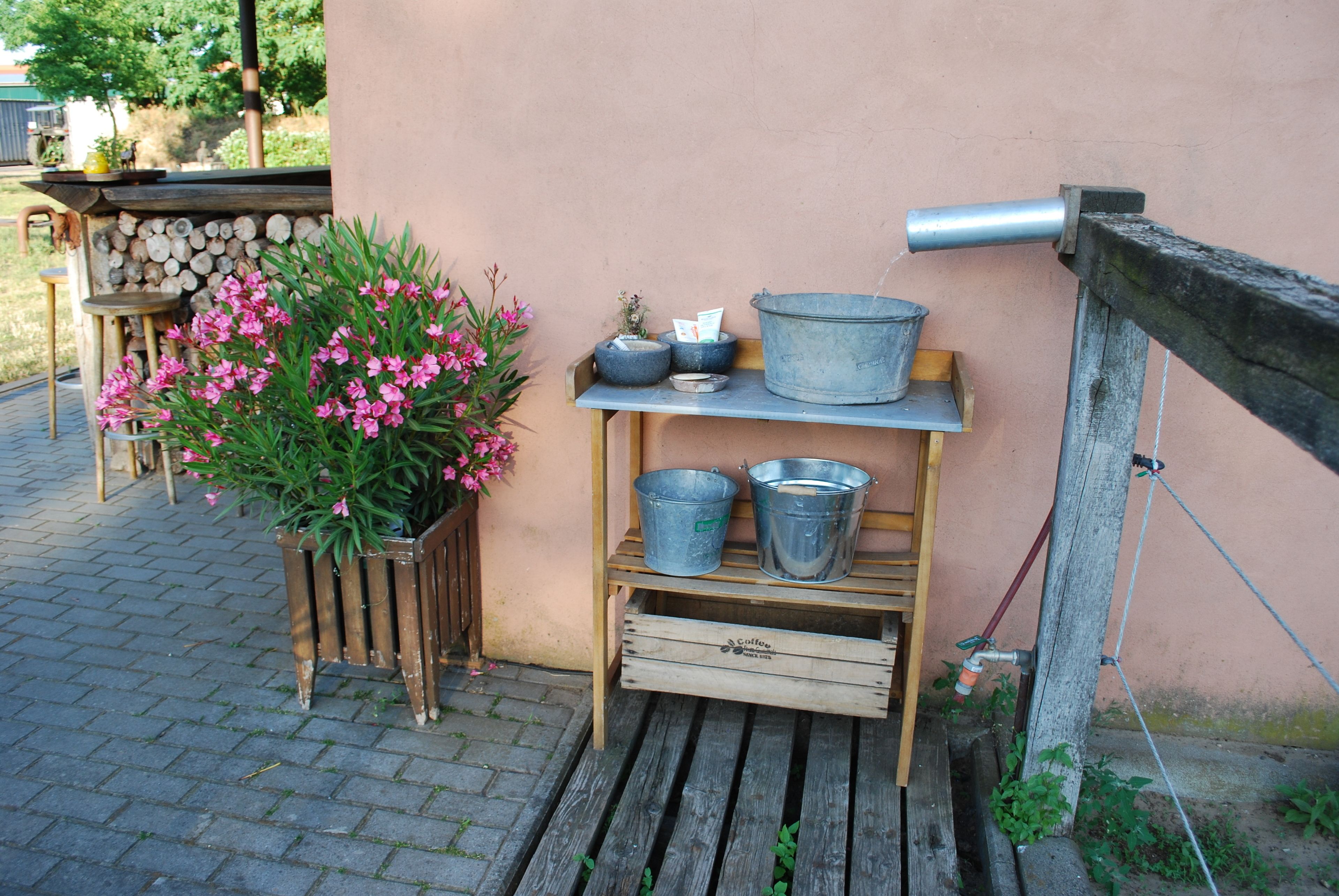 A rustic outdoor setting with a wooden bench, metal buckets, and a potted plant with pink flowers.