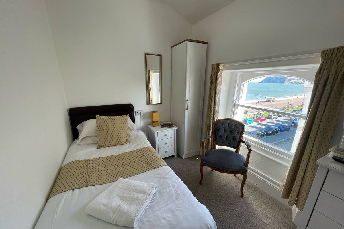 A hotel room with a bed, a chair, a bedside table, a mirror, and a window with a view of the beach and cars.