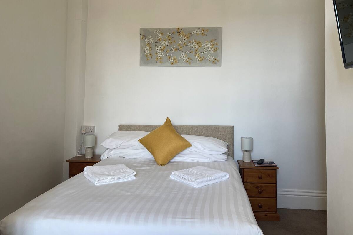 A neatly made bed with white linens, a mustard-colored pillow, and two folded towels on the side. There is a nightstand with a lamp and a power outlet on the left. A painting hangs on the wall above the bed.
