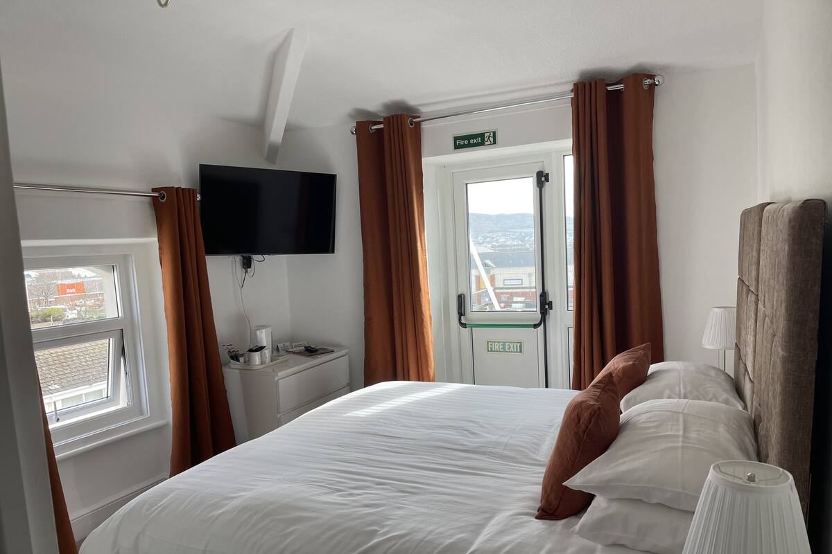 A hotel room with a bed, a TV mounted on the wall, a bedside table with a lamp, and a view of the outside through a window.