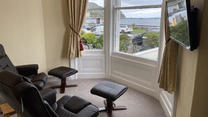 A room with a large window, a black leather sofa, a small ottoman, and a mounted TV. - The Shelbourne (Llandudno)