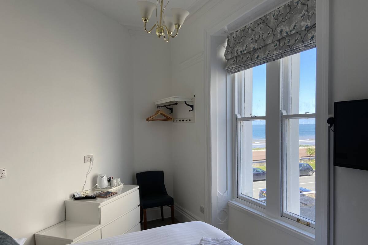 A hotel room with a bed, a desk, a chair, a hanging coat rack, and a window with a view of the beach and ocean.