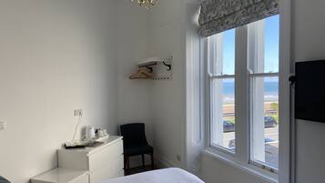 A hotel room with a bed, a desk, a chair, a hanging coat rack, and a window with a view of the beach and ocean.