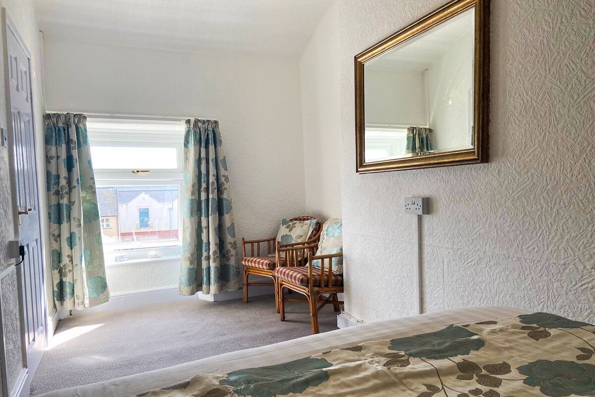 A room with a bed, a chair, a window with curtains, and a wall-mounted mirror.