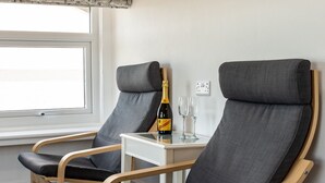 Two grey lounge chairs with wooden frames, a small white table with a bottle and two glasses, and a framed painting on the wall. - The Shelbourne (Llandudno)