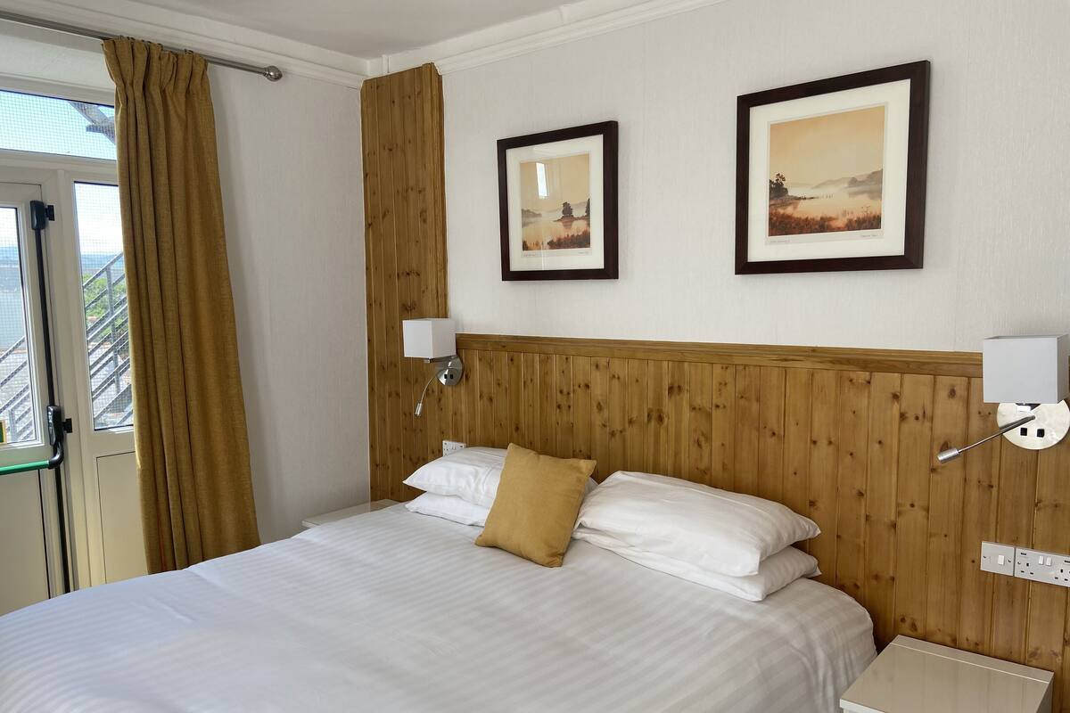 A neatly made bed with white linens and a single tan pillow, a wooden headboard, two wall-mounted reading lamps, and two framed landscape paintings.