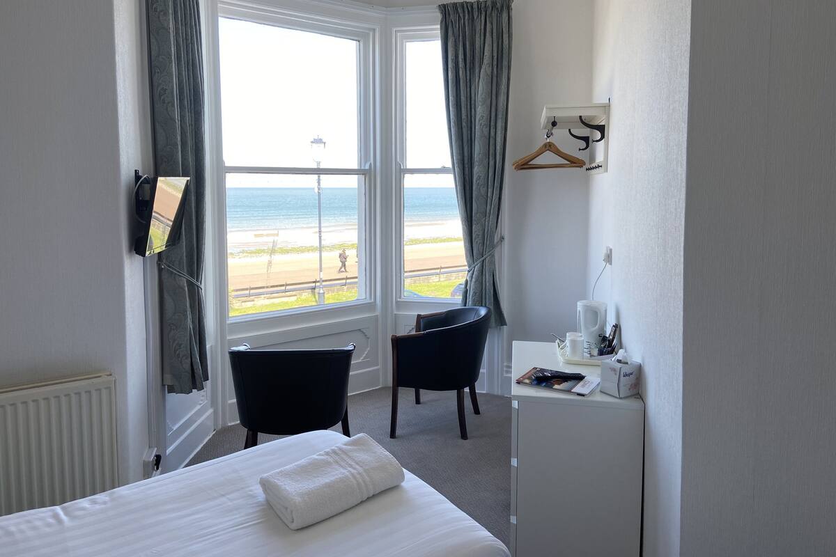 A hotel room with a bed, a desk with a chair, a small table with a coffee maker, and a view of the beach.