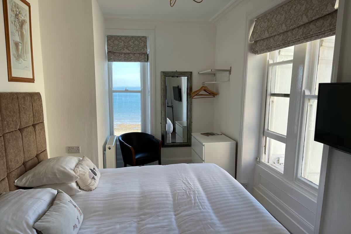 A bedroom with a bed, a chair, a bedside table, a mirror, and a TV.