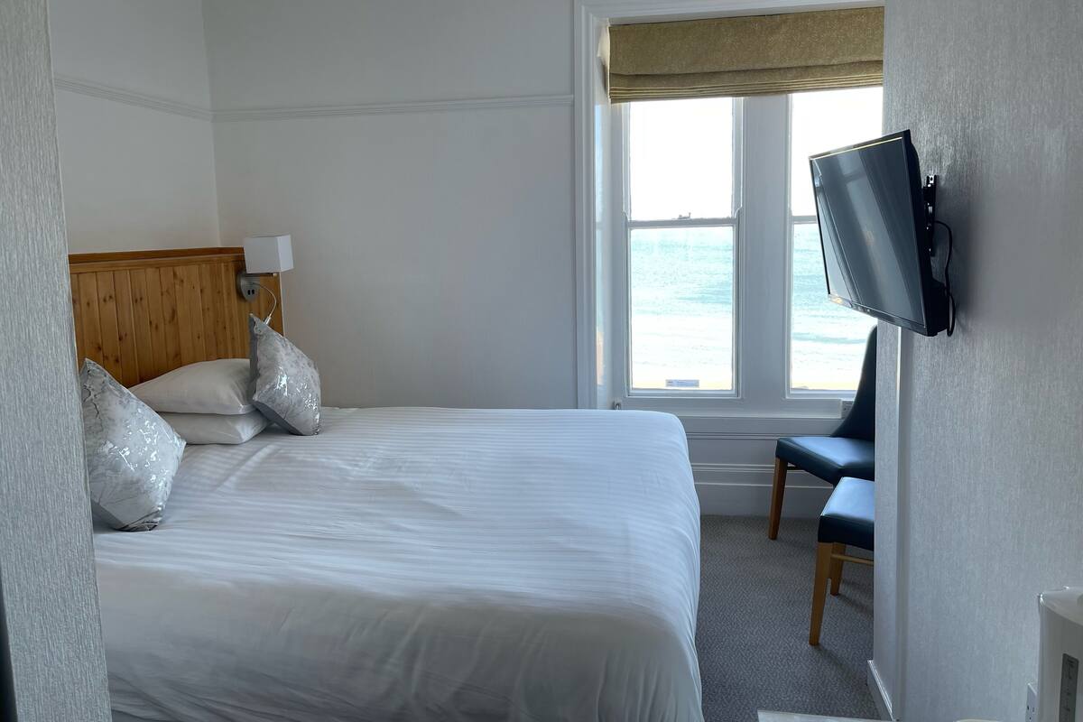 A hotel room with a bed, a television mounted on the wall, a chair, and a window with a view of the sea.