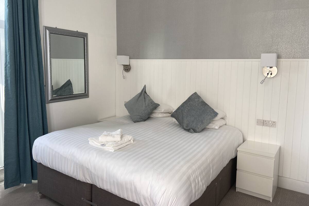 A neatly made bed with pillows, a bedside table, a nightstand, a wall-mounted light, and a mirror.