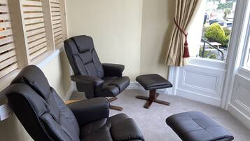 A room with two black leather chairs and a matching ottoman, a wooden slatted window, and a view of a parking lot through a curtain.