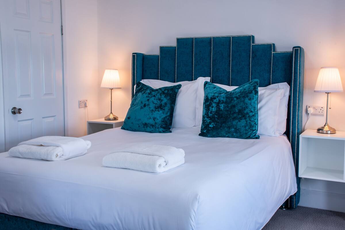 A neatly made bed with white linens and teal pillows, two bedside lamps, and a white nightstand.