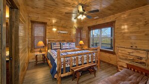 Un dormitorio de cabaña de madera con una cama de madera, dos lámparas de noche, un ventilador de techo y un ventanal grande.
