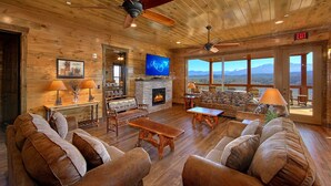 Amplia sala de estar con pisos de madera, chimenea de piedra, televisor y un ventanal con vista a la montaña.