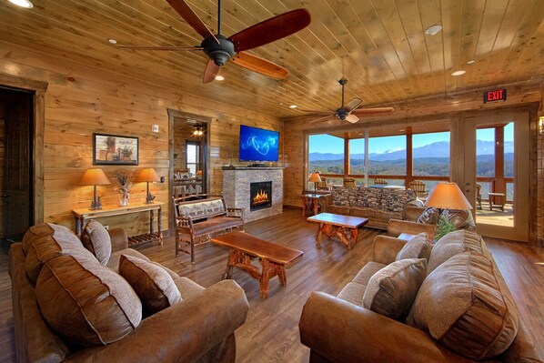 A spacious living room with wooden floors, a stone fireplace, a television, and a large window offering a mountain view.