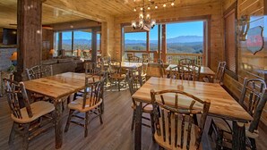 Un interior rústico de madera con varias mesas y sillas para comer, amplios ventanales que ofrecen vistas a las montañas y una zona de estar acogedora con chimenea.