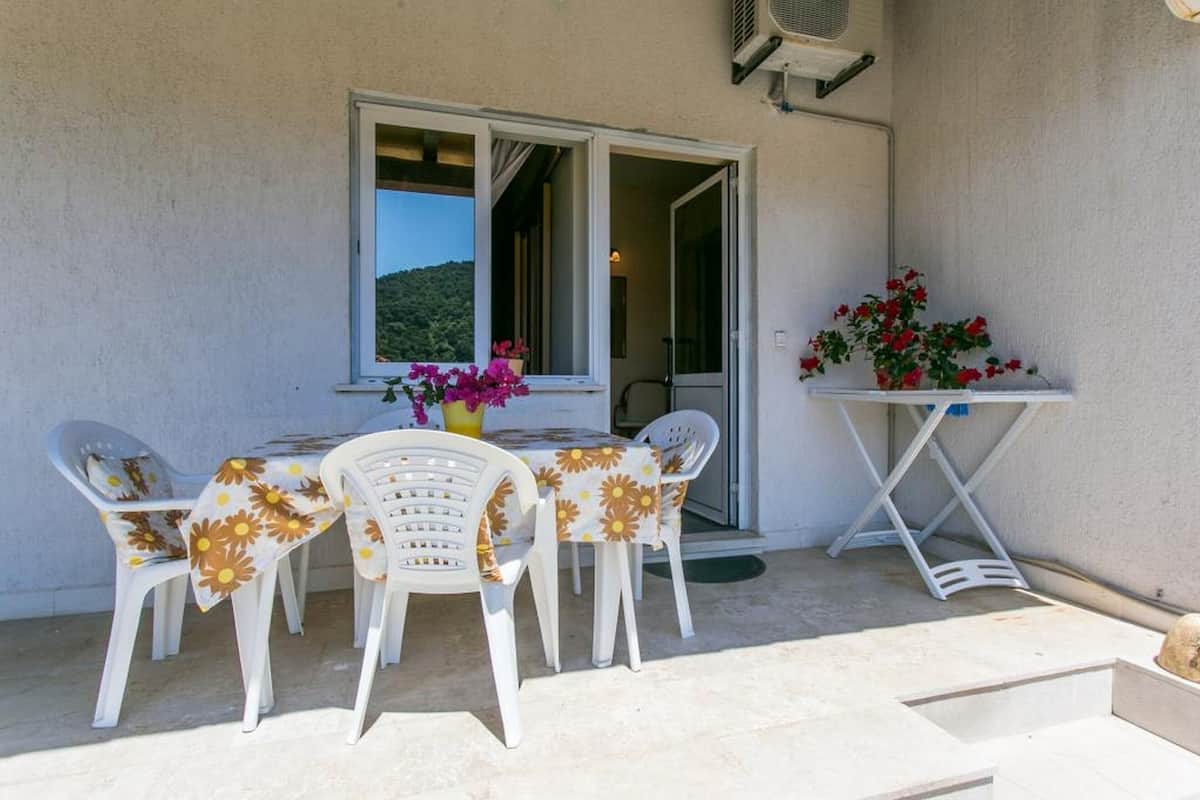 An outdoor seating area with a table and chairs, a window with a view of a hill, and a small table with flowers.