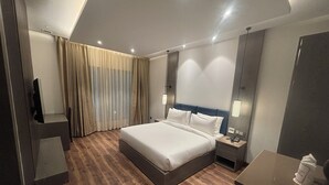 Une chambre d’hôtel moderne avec un grand lit, un plancher en bois, une table de chevet avec une lampe, un miroir et une fenêtre avec des rideaux.