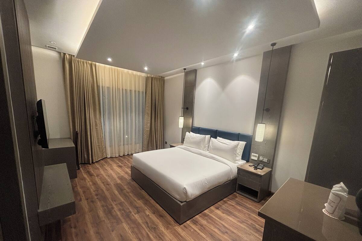 Ein modernes Hotelzimmer mit einem großen Bett, Holzboden, einem Nachttisch mit Lampe, einem Spiegel und einem fenster mit Vorhängen.