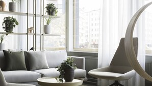 A modern living room with a grey sofa, a round coffee table, and a white armchair. There are potted plants on shelves and a large window with curtains. - Noli Sörnäinen (Helsinki)