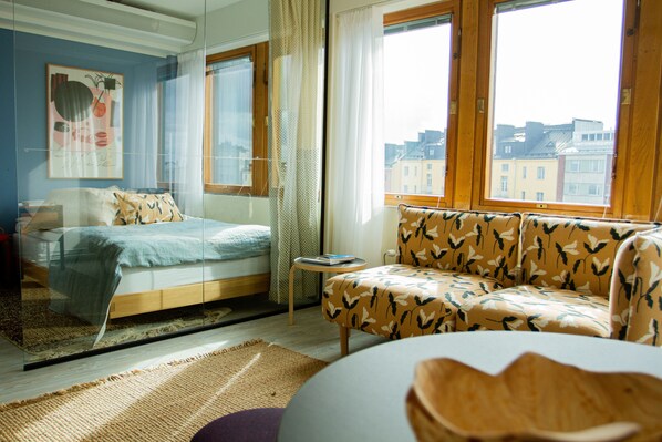 A modern bedroom with a bed, a sofa, and a view of buildings through large windows. - Noli Sörnäinen (Helsinki)