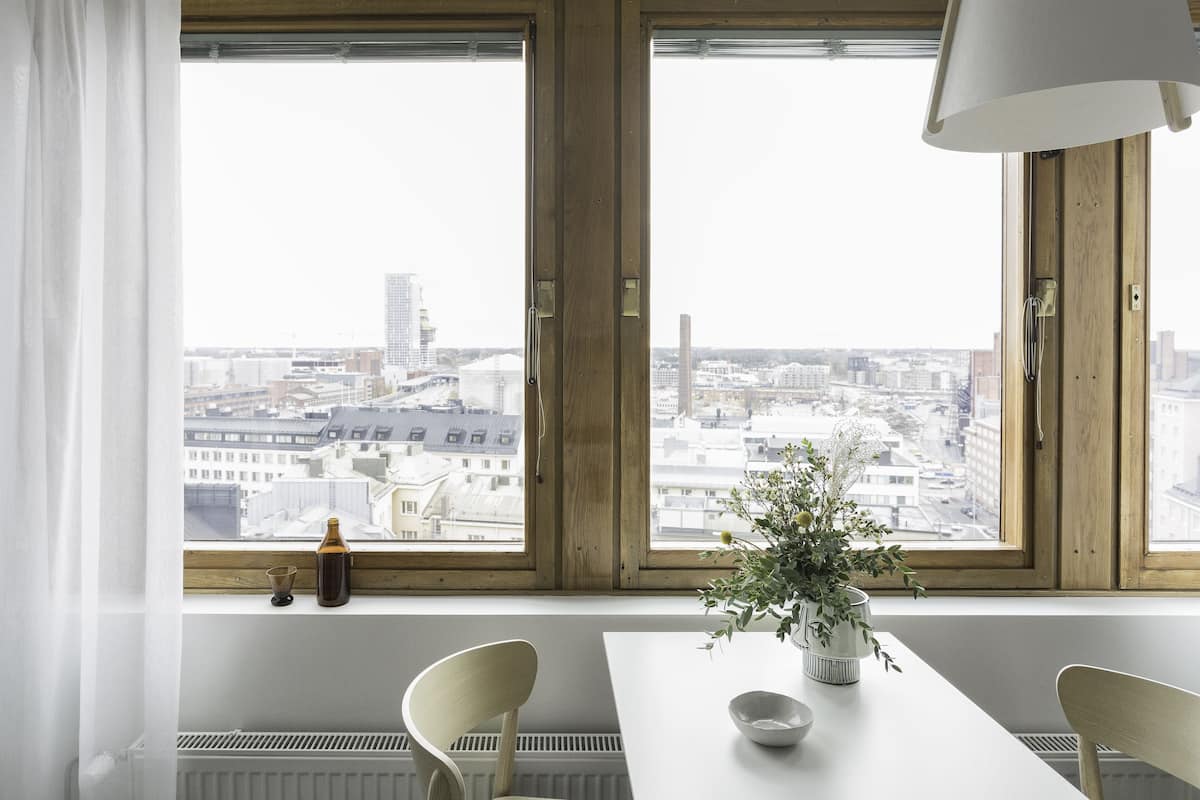 Une pièce avec une grande fenêtre, une table blanche, deux chaises, une lampe et une plante en pot.
