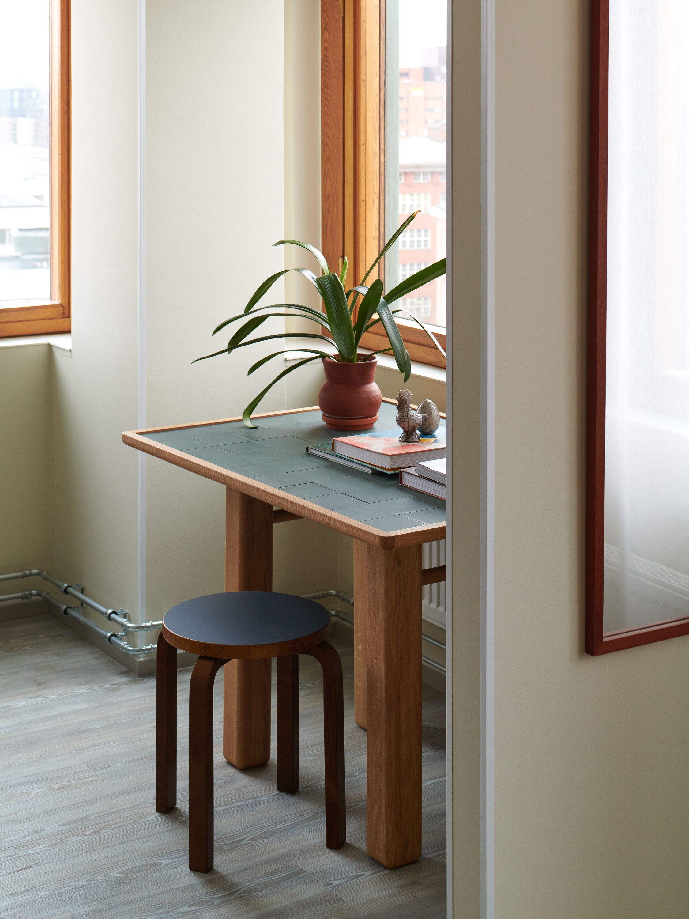 A corner desk with a potted plant and a small figurine, a stool, and a framed picture on the wall.