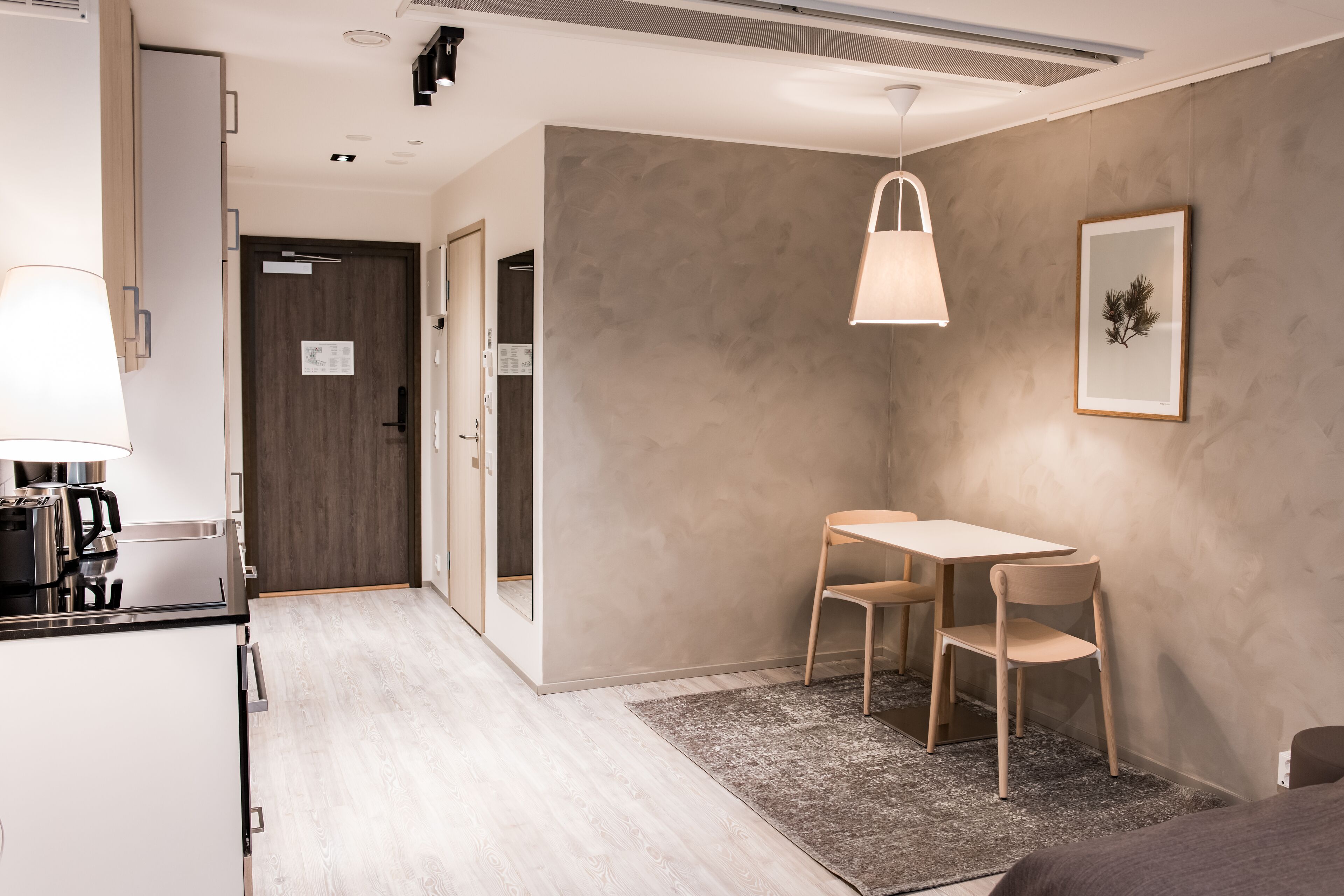 A modern hotel room with a kitchenette, a dining area with a table and chairs, a framed picture on the wall, and a ceiling light fixture.