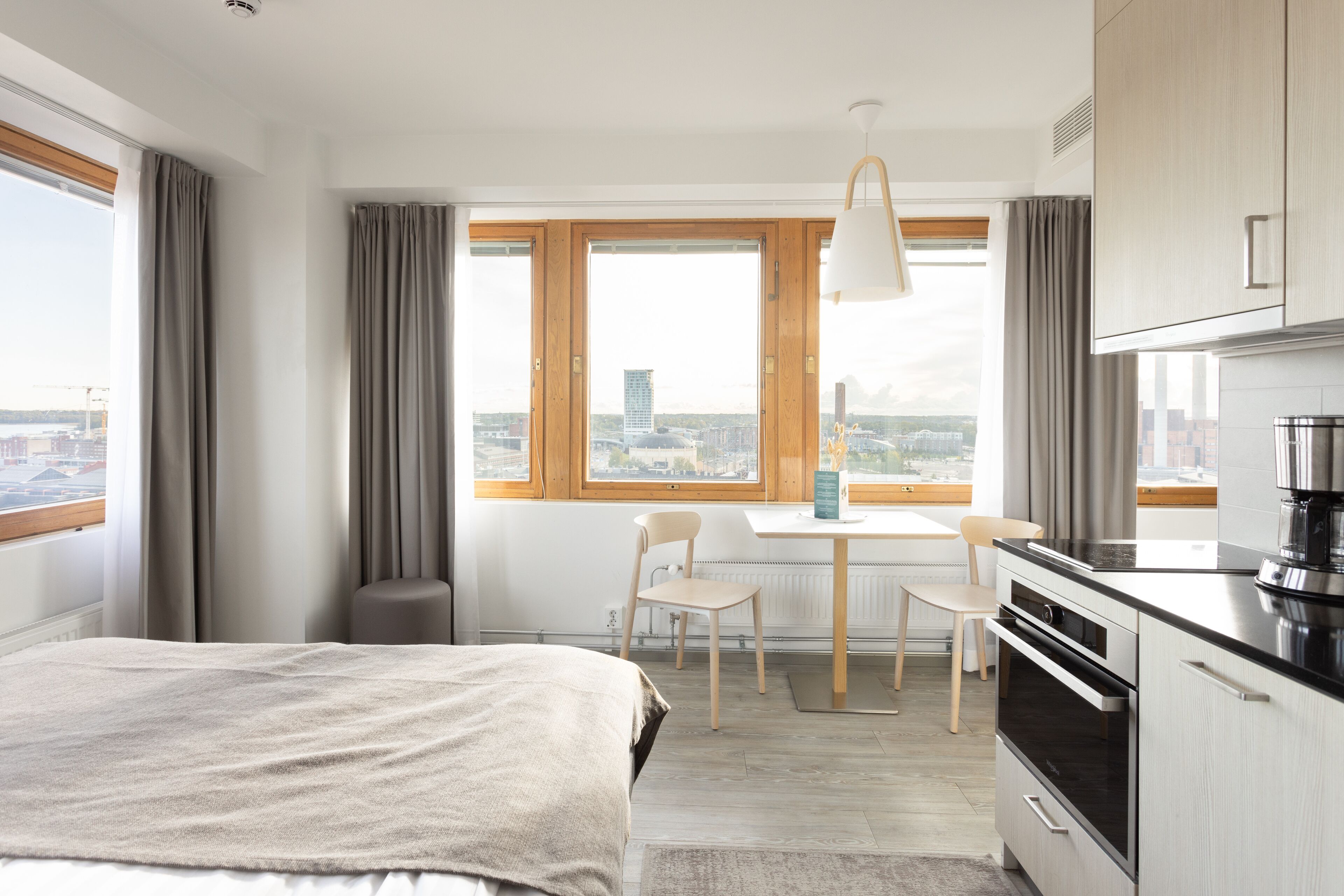 A modern hotel room with a bed, a dining area with chairs, a kitchen with a stove, and large windows offering a city view.