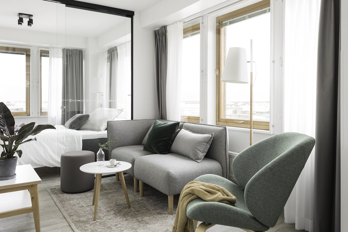 Une chambre d’hôtel moderne avec un canapé gris, un fauteuil vert, une petite table ronde et une plante en pot.