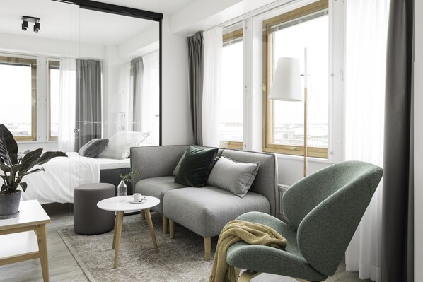 Habitación de hotel moderna con sofá gris, sillón verde, una mesita redonda y una planta en maceta.
