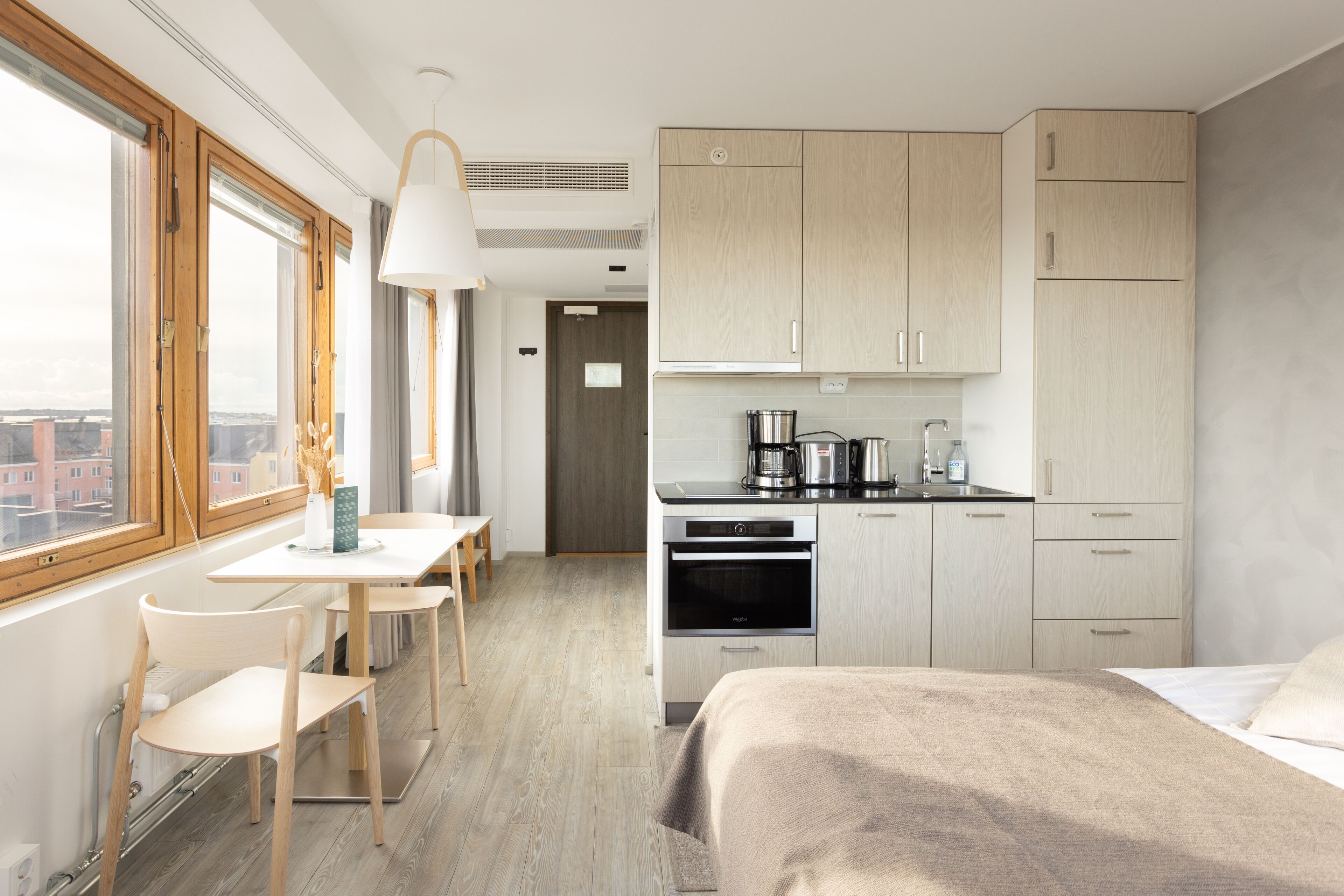 A modern hotel room with a bed, a desk, a chair, a kitchenette, and a large window offering a city view.