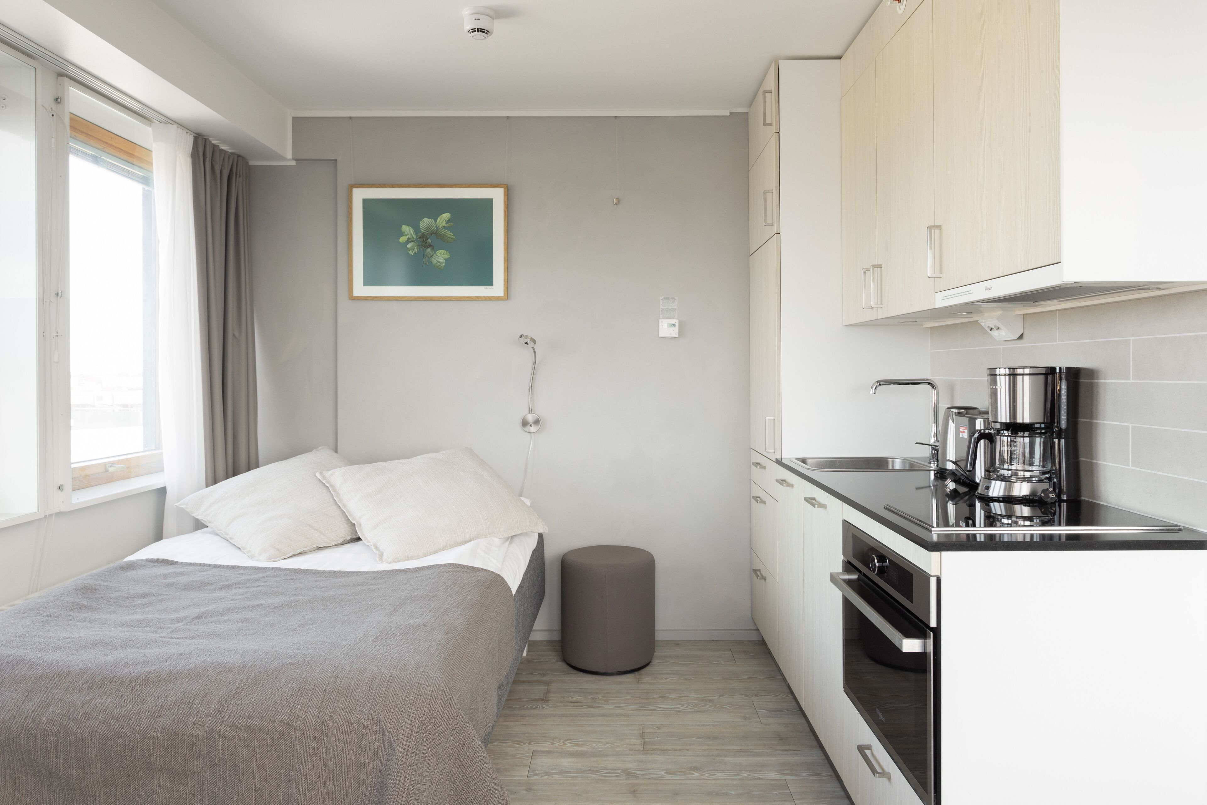 A compact bedroom with a bed, bedside table, and a small stool. Adjacent is a kitchenette with a stove, sink, and coffee maker. A framed picture hangs on the wall.