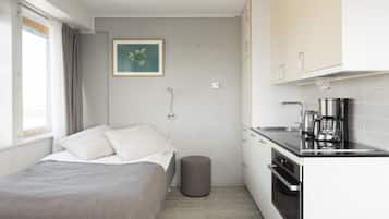 A compact bedroom with a bed, bedside table, and a small stool. Adjacent is a kitchenette with a stove, sink, and coffee maker. A framed picture hangs on the wall.